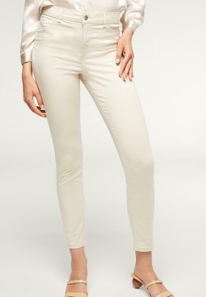 Pantaloni slim fit color crema indossati con sandali beige con tacco basso e camicetta in raso rosa chiaro a maniche lunghe, in piedi contro uno sfondo semplice.