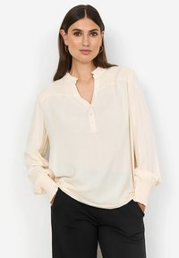 Light beige blouse with a ruffled neckline, buttoned front, and elastic cuffs. Smooth fabric, relaxed fit, paired with black trousers.