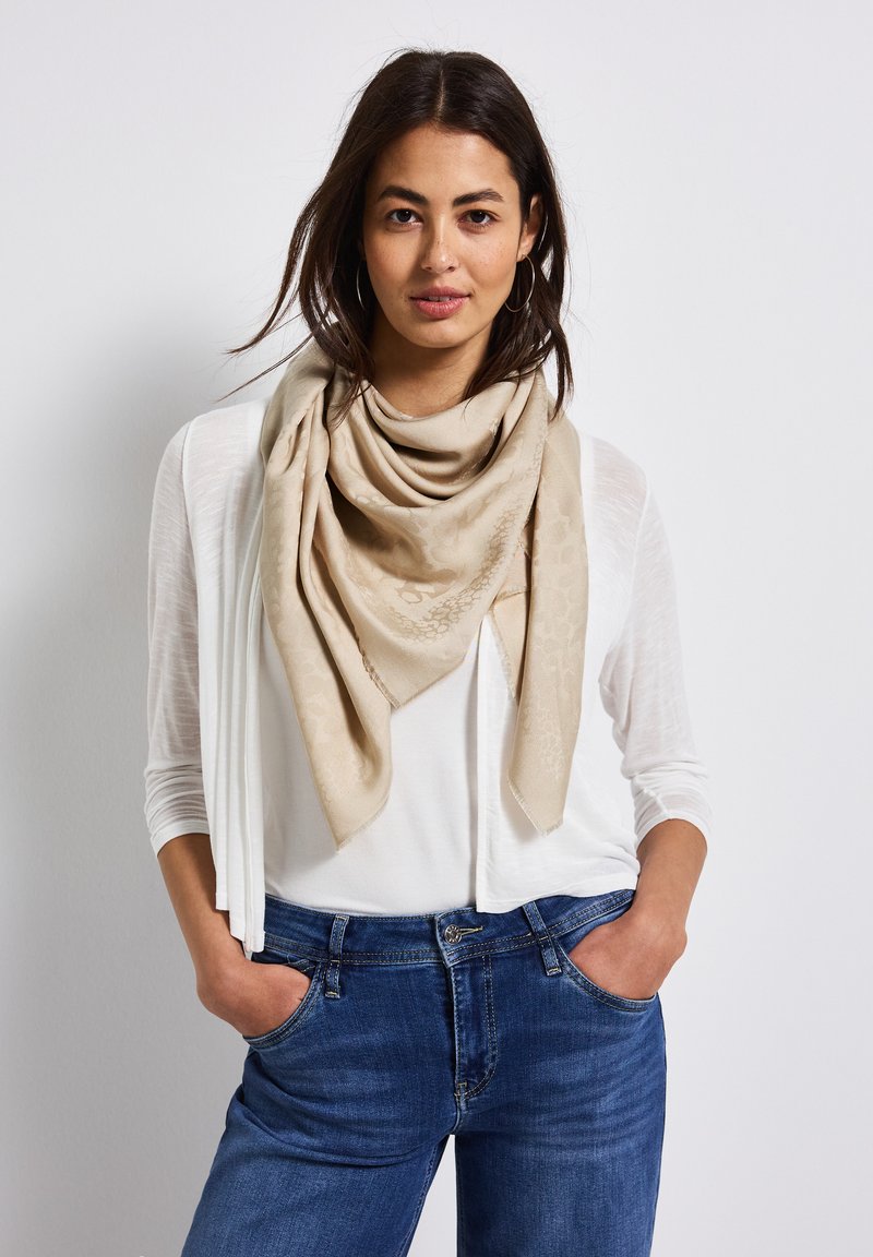 Beige sjaal met een subtiel patroon, om de hals gedrapeerd. Draag over een witte top en blauwe jeans, die een casual outfit toont.