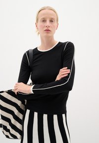 Melna garām piedurknēm kreklā ar baltu apdari, saskaņota ar melnu un balti svītrainu svārku. Nēsā svītrainu somu ar divu toņu rakstu.