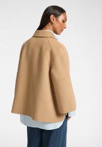 Beige ullkappa med hög krage, vida ärmar och rena linjer. Buren över en ljusblå skjorta, i kombination med denim.