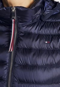 Laivastonsininen toppatakki, jossa on kiiltävä pinta, vaakasuoria tikattuja osioita, korkea kaulus ja hopeanvärinen vetoketju, jossa on punavalkoinen vetimetaltta.