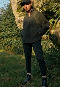 Donkergroen fleece trui met een half ritssluiting en voorzakken, gecombineerd met zwarte camouflage leggings en zwarte enkellaarsjes met grijze accenten.