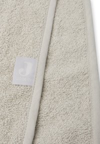 Serviette en coton beige clair, présentant une surface texturée. Bordée d'une finition lisse. Comprend une petite étiquette de logo dans le coin.