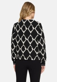 Vrouw met blond haar, gekleed in een zwart-wit diamantpatroon trui en zwarte broek, gezien van achteren tegen een effen achtergrond.