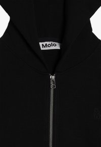 Sudadera con cremallera negra que presenta una cremallera plateada, una capucha grande y una tela texturizada con un logo embosado cerca de la cremallera.