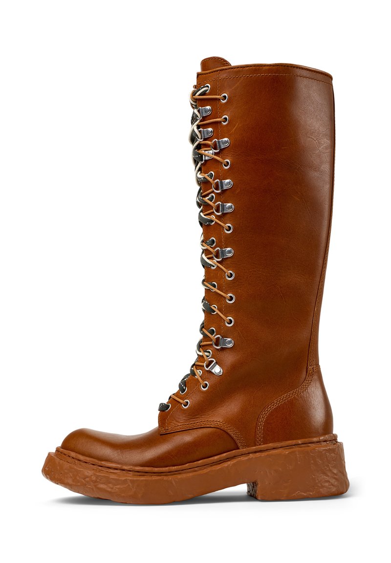 Bota de cuero marrón hasta la rodilla con punta redondeada, ojales plateados y cordones negros. Suela gruesa y texturizada con un tacón ligeramente elevado.