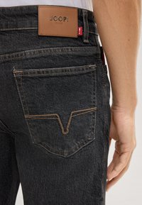 Jeans di denim nero con un'etichetta in pelle che riporta "JOOP! JEANS", cuciture marroni a contrasto e un distintivo design delle tasche a forma di V.