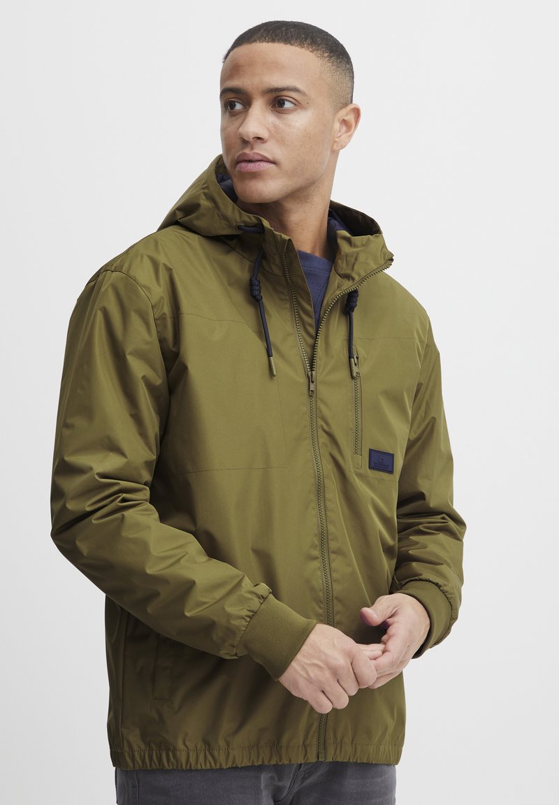 Olive green hooded jacket made from a lightweight material; features zip front, elastic cuffs, and a small logo patch on the chest.