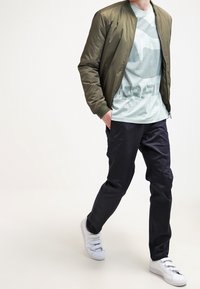 Man som bär en grön bomberjacka, grå grafisk t-shirt, svarta byxor och vita sneakers, går med handen i fickan.