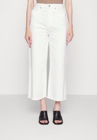 Jeans bianchi a gamba larga con orlo sfrangiato, realizzati in denim. Abbinati a sandali modello slide in pelle nera. Sfondo semplice, focus sul capo inferiore.