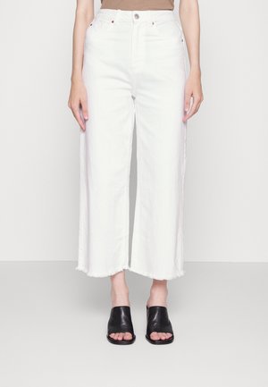 Jeans bianchi a gamba larga con orlo sfrangiato, realizzati in denim. Abbinati a sandali modello slide in pelle nera. Sfondo semplice, focus sul capo inferiore.