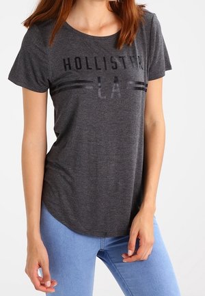Camiseta de manga corta gris oscuro con la impresión en negro "HOLLISTER LA". Con dobladillo redondeado, tejido suave y corte relajado, combinada con jeans de color azul claro.
