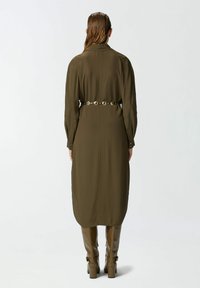 Maslinasto zelena haljina ravnog kroja, dugih rukava i kragne. Ima zlatni pojas s kružnim detaljima. Glatka tkanina s mekanom teksturom.