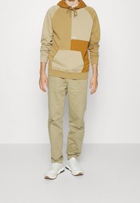 Sweat à capuche ton sur ton en marron et beige avec une poche avant, associé à un pantalon beige clair et des baskets blanches avec une semelle en gomme.