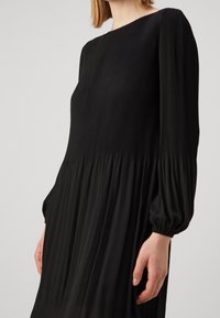 Robe noire midi avec de longues manches bouffantes. Présente un design plissé et un corsage ajusté qui se transforme en une jupe fluide. Texture lisse.