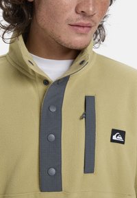 Fleece pullover in lichtgeel met grijze accenten, voorzien van een hoge kraag, drukknoopjes en een ritszak aan de voorkant.