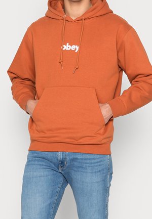 Personne portant un sweat à capuche orange rouille avec le logo blanc "obey", mains dans la poche avant, associée à un jean bleu.