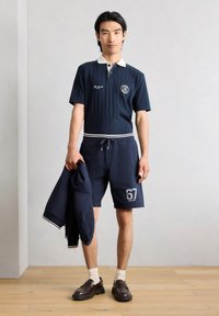 Homme portant un polo bleu marine et un short avec le numéro 67, tenant une veste bleu marine assortie, debout sur un sol en bois contre un mur uni.
