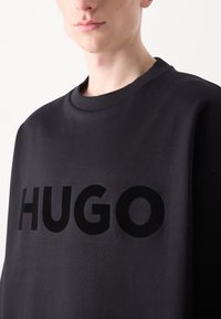 Jeune personne portant un sweat-shirt noir avec les lettres larges et audacieuses "HUGO" sur la poitrine, sur un fond blanc uni.