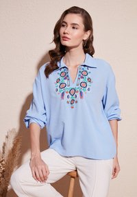 Hellbluse mit langen Ärmeln und Kragen, die um den Ausschnitt herum mit filigraner floralem Stickerei in Rot, Pink und Blau verziert ist. Weicher Stoff.