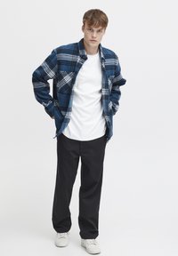 Blauwe geruite shirt met witte en zwarte accenten, gedragen over een wit T-shirt; gecombineerd met zwarte broek en witte sneakers.