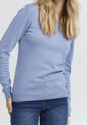 Blonde Frau trägt hellblauen Pullover mit langen Ärmeln und dunkelblaue Jeans, steht vor hellem, einfarbigem Hintergrund.