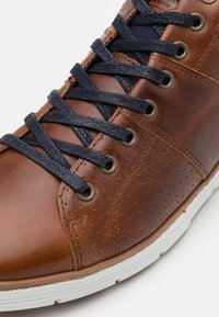 Hnědé kožené boty s námořnickými šněrovadly, které mají perforované detaily a kulaté kovové očko. Bílá gumová podrážka doplňuje design.