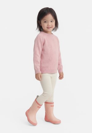 Jeune fille souriante, portant un pull rose avec des motifs de cœurs, des leggings crème et des bottes de pluie roses, debout sur un fond clair.
