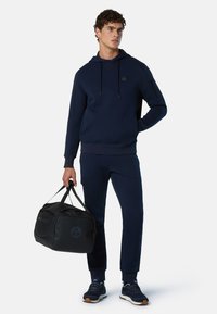Sudadera y pantalones marinos de tela suave, con capucha y bolsillo con cremallera. Bolsa de deporte negra con logo y zapatillas marinas con detalles en marrón.