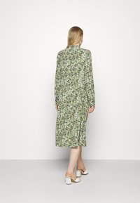 Zelené květinové midi šaty s dlouhými rukávy, staženým pasem, volnou sukní a vysokým límcem. Modelka nosí bílé muly s blokovým podpatkem.