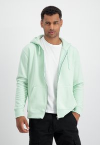 Uomo con una felpa verde chiaro con zip sopra una maglietta bianca e pantaloni neri, in piedi contro uno sfondo chiaro e semplice.