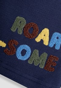 Mornarski tekstil s kolorističnim, teksturiranim vezom koji ispisuje "ROAR-SOME" u zelenim, smeškim, plavim i žutim slovima.