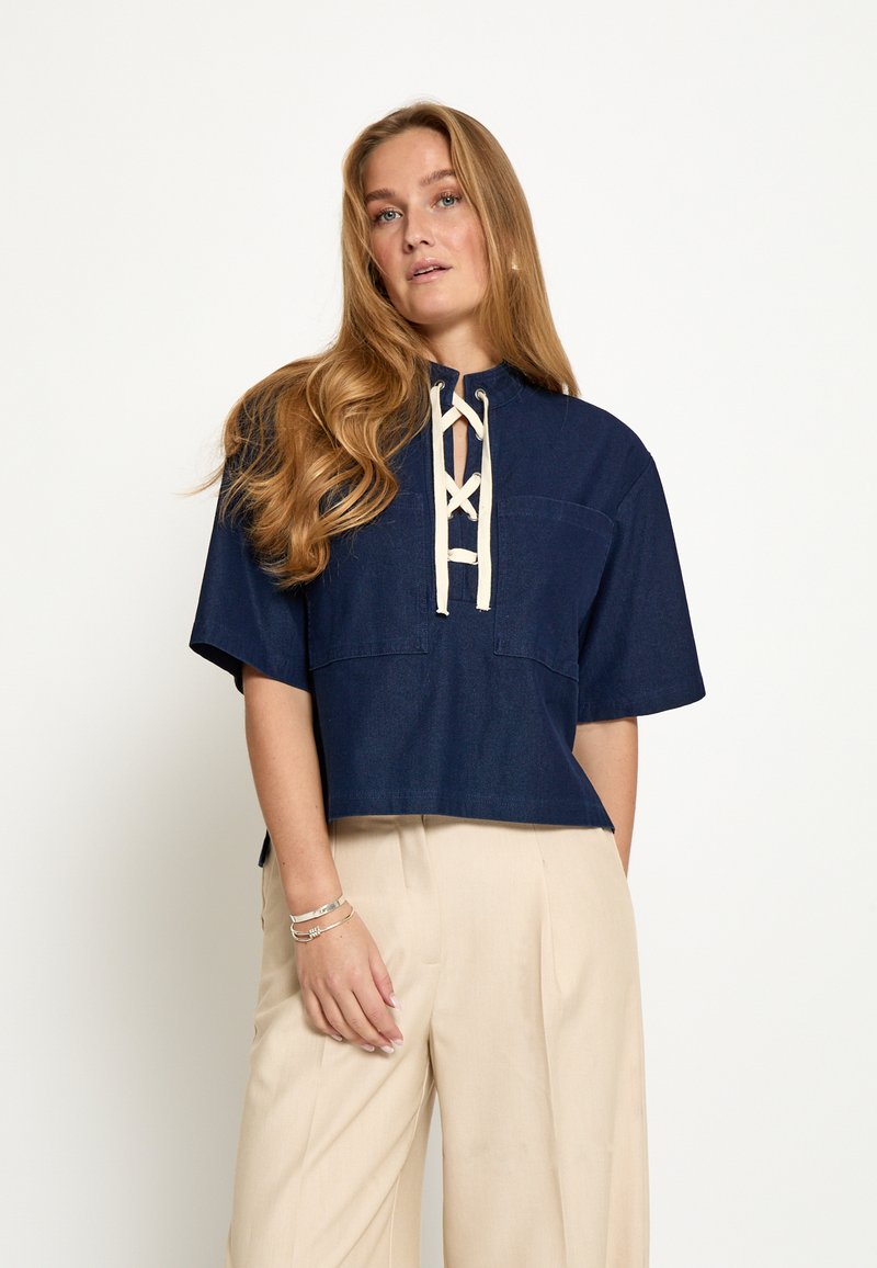 Woman with long hair wearing a navy blue lace-up short-sleeve top and beige wide-leg pants, standing against a plain white background.