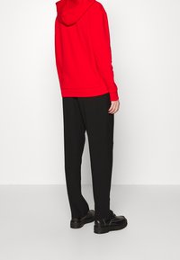 Červená mikina s kapucí ve volném střihu, doplněná volnými černými kalhotami, viděná ze zadní strany. Jednoduchý design a hladká textura.