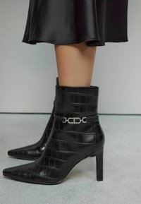 Bottines noires avec un bout pointu, en cuir gaufré crocodile. Présentent une accent en chaîne argentée autour de la cheville et un talon aiguille.