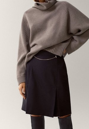 Persoon draagt een grijze coltrui, ingestopt in een zwarte rok met een dun kettinkdetail, gecombineerd met zwarte kniehoge laarzen.