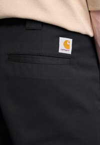 Sorte bukser lavet af et slidstærkt stof med en baglomme og et hvidt mærke med et orange logo. Glat tekstur, klassisk design.