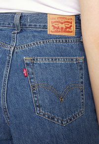 Jeans de ganga azul com um patch com o rótulo "Levi Strauss & Co.", um design clássico de bolso traseiro com costura amarela e uma etiqueta de logo vermelha.