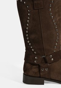 Botte en daim marron avec des accents de clous argentés sur le côté, une sangle bouclée et un talon bas et rectangulaire.