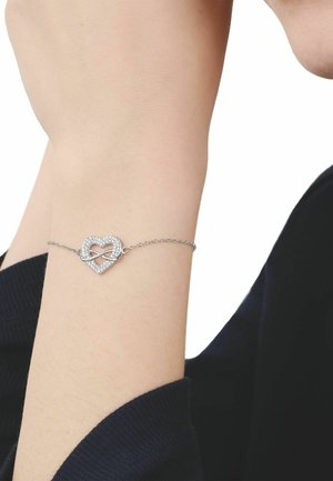 Pulsera delicada de plata con un colgante en forma de corazón infinito adornado con pequeños cristales, en una muñeca que lleva una prenda de manga larga oscura.