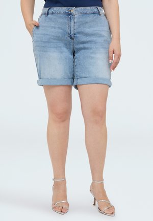 Person iført lyseblå denimshorts med opkrogede kanter og små rhinsten, navy top og sølvfarvede højhælede sandaler, står med afslappede hænder.