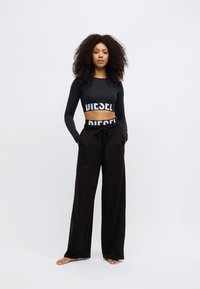 Melns garroku kultūraugs ar baltu "DIESEL" logotipu, savienots ar vaļīgām melnām biksēm ar aukliņu viduklī un kabatām.