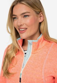 Giacca in fleece di un vivace colore corallo con collo con zip, dettagli in azzurro chiaro e tessuto texturizzato. Presenta un dettaglio con logo sulla zip.