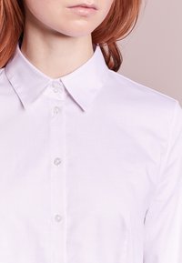 Camicia bianca con colletto, caratterizzata da un motivo testurizzato, con bottoni visibili e una vestibilità su misura, che enfatizza linee pulite e un tessuto morbido.