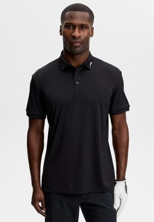 Mand iført en sort kortærmet poloshirt og en hvid golfhandske på venstre hånd, stående foran en ensfarvet baggrund.