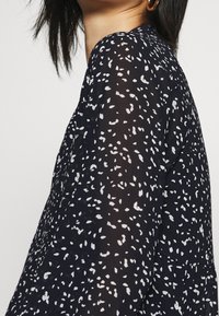 Blusa a fantasia bianco e nero con un tessuto trasparente e testurizzato; presenta un collo rotondo e maniche fluide. Gli accenti includono forme astratte irregolari.