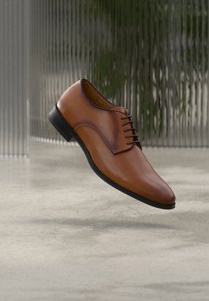 Brązowy skórzany męski but wizytowy na sznurowadła, unoszący się nad polerowaną betonową podłogą, na tle teksturowanej szklanej ściany.