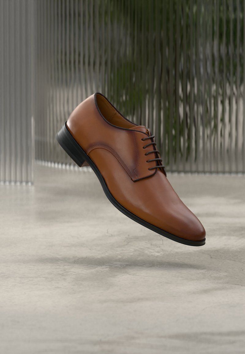Καφέ δερμάτινο παπούτσι επίσημο με κορδόνια, αιωρούμενο πάνω από γυαλισμένο τσιμεντένιο πάτωμα με φόντο υαλωμένη υφή γυαλιού.