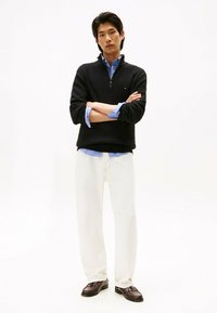 Schwarzer Strickzipper, darunter ein blau gestreiftes Hemd, cremefarbene Hose und dunkle Slipper. Das Model steht mit verschränkten Armen vor einem schlichten Hintergrund.